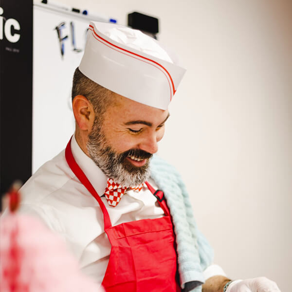 a chef wearing a white chef outfit with red strips and a red apron and smiling