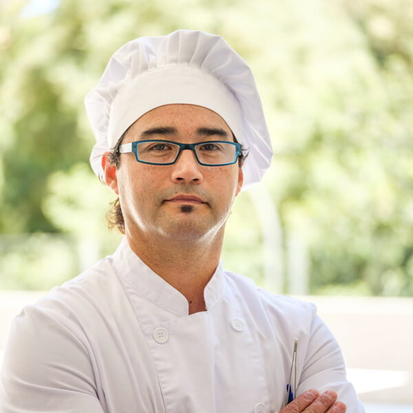 a chef wearing a white chef outfit with red strips and a red apron and smiling
